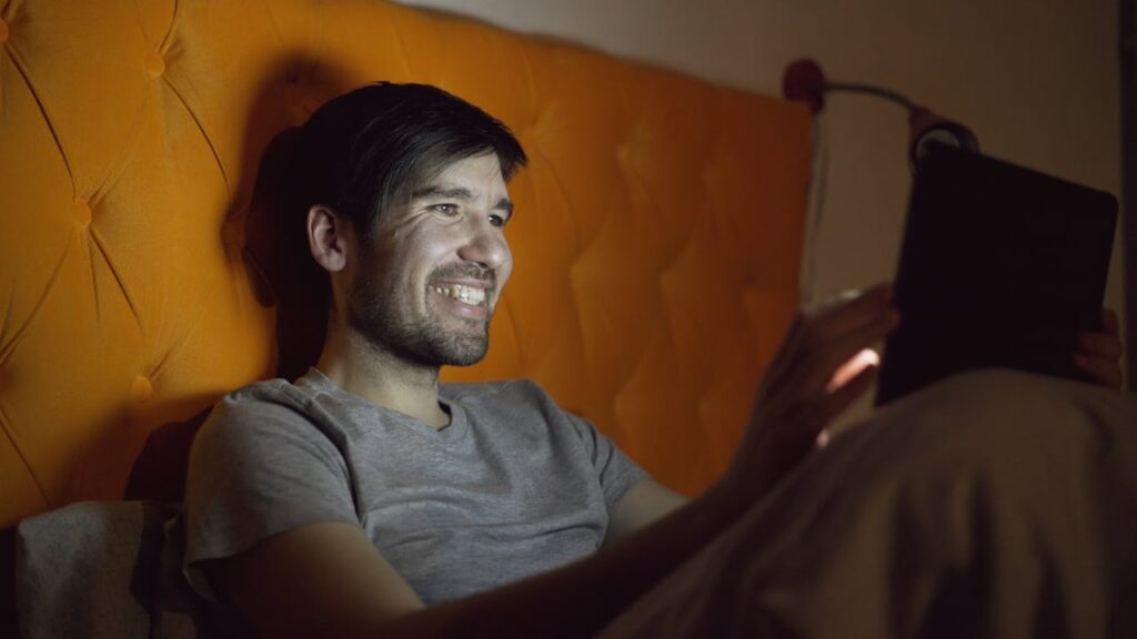 A man relaxes in bed at night, illuminated by his tablet screen.