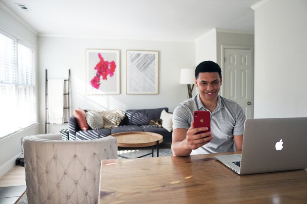 Man sitting in a modern living room, using a smartphone and laptop, showcasing remote work lifestyle.