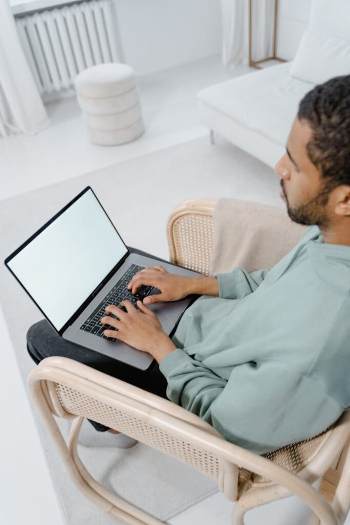 A man sits in a rattan chair typing on a laptop in a bright, modern home setting.